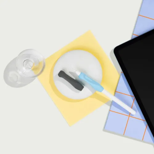 Penngrepp för Apple Pencil visas: blått grepp på pennan och kolsvart grepp bredvid, på papper intill surfplatta och glas