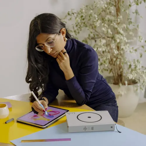 Blått silikonpenngrepp på Apple Pencil som hålls över en iPad, med produktlåda bredvid på bordet och papper i starka färger.