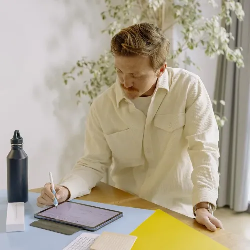 Paperlike Pencil Grips V2 på Apple Pencil, blått grepp i användning på surfplatta; person lutar sig över bord med tillbehör.