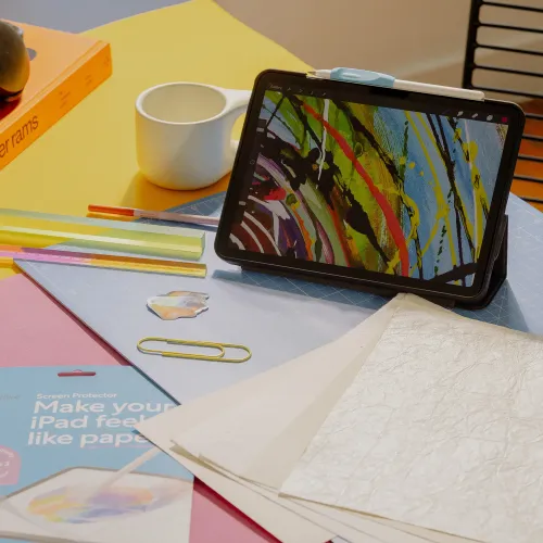Paperlike 3-skärmskyddets förpackning på skrivbord bredvid iPad i fodral med penna, linjaler, papper, gem och en vit mugg.