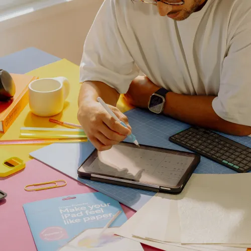 Paperlike 3 screen protector on iPad with matte finish when one hand draws with stylus; packaging and tools are on the table next to it.